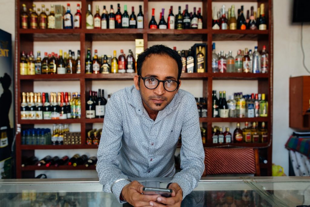 Man In Eyeglasses Holding A Phone Behind The Glass Counter Of A Liquor Store
